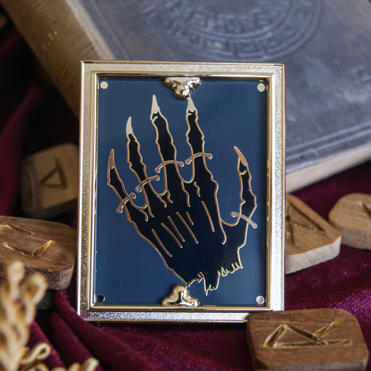 Photo of an enamel pin depicting a mummified hand strapped down like a museum exhibit. Around the hand is a 3D golden frame, and behind the pin is a book and some scattered wooden runes.