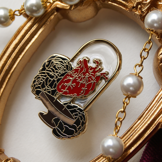 Enamel pin depicting a human heart inside a bell jar, backed by black peonies. Around the pin are pearls and a gold frame.