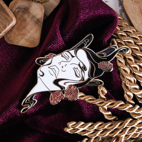 Enamel pin depicting the Shakespeare character Ophelia after her drowning. Her dark hair surrounds her face and red flowers are tangled in it.