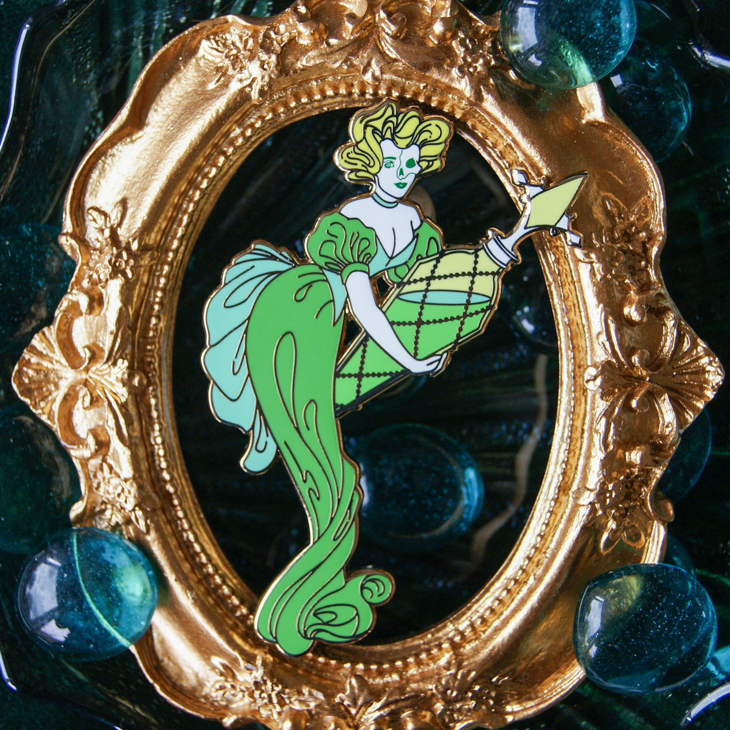 Photo of an enamel pin featuring a victorian woman in a green dress, holding a huge vial of green liquid - absinthe. 