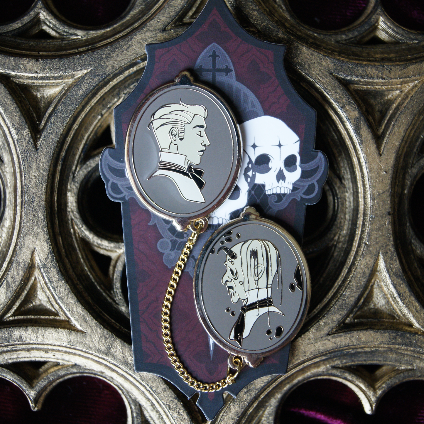 Photograph of two enamel pins connected by a chain. They depict a young Dorian Grey on one pin, and a rotted corpse on the other. Behind the pin is a coffin shaped backing card, and a gothic carved window. 