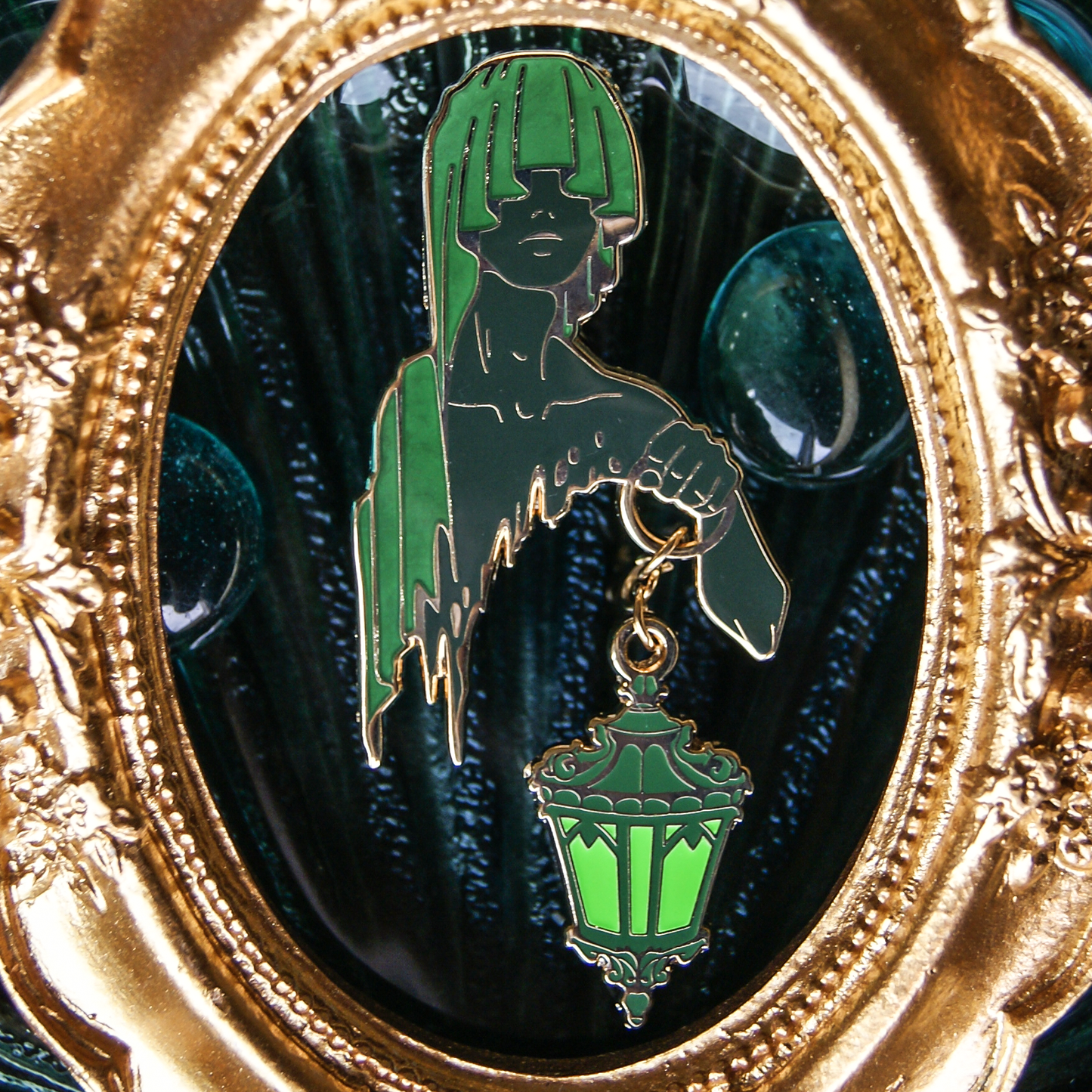 An enamel pin depicting a hooded figure holding an ornate lantern on a chain. Their cloak and skin are green. Behind is an oval, golden frame. 