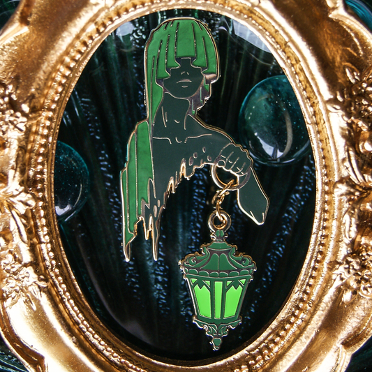 An enamel pin depicting a hooded figure holding an ornate lantern on a chain. Their cloak and skin are green. Behind is an oval, golden frame. 