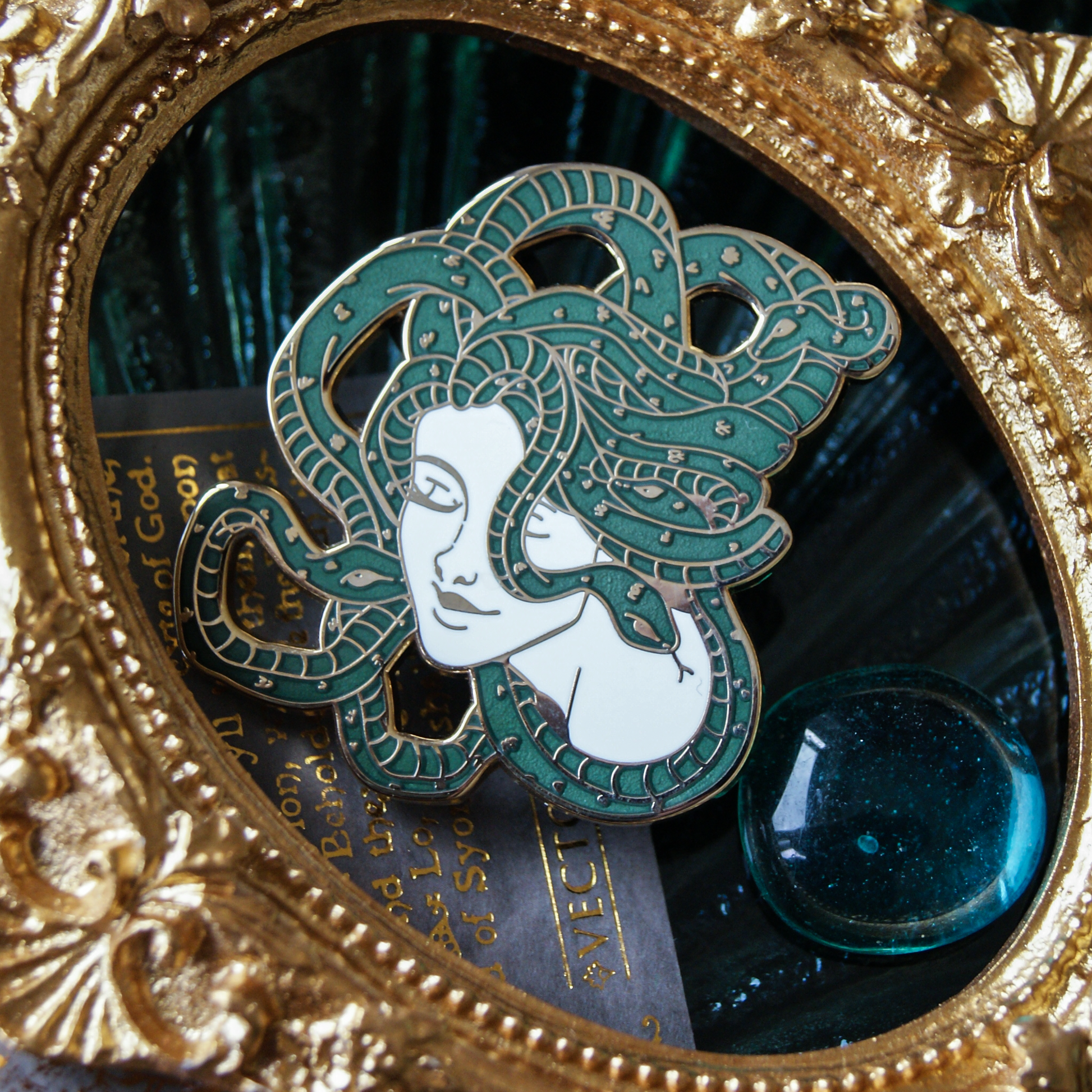 Enamel pin depicting Medusa; a woman with a head of snakes. One snake is kissing her on the cheek. A golden frame surrounds them. 