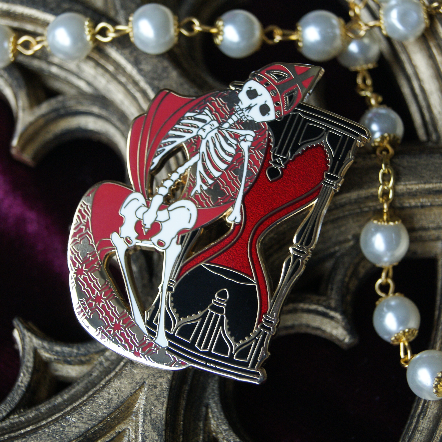 Enamel pin depicting a skeleton in vestal robes backed by a red sandglass timer. Behind the pin are pearls and a gothic window carving. 