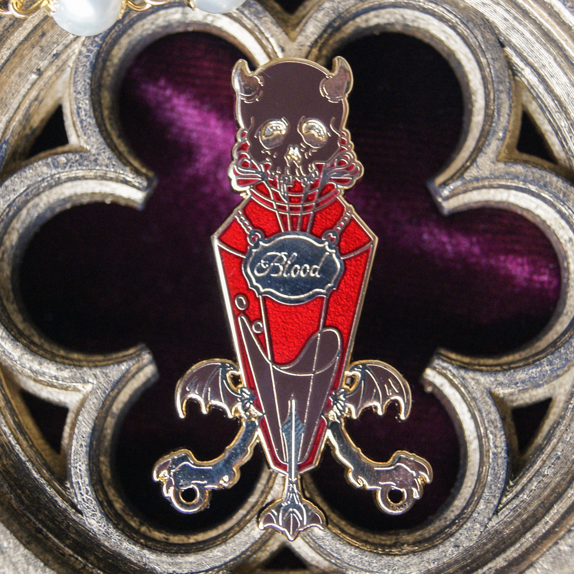 Enamel pin depicting a vial labelled 'blood'. Inside is a red liquid. The background is a carving of a gothic window. 