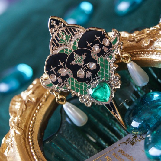 Photograph of a pin featuring three skulls above a dagger, matching tear pearls, and a heart gem. 