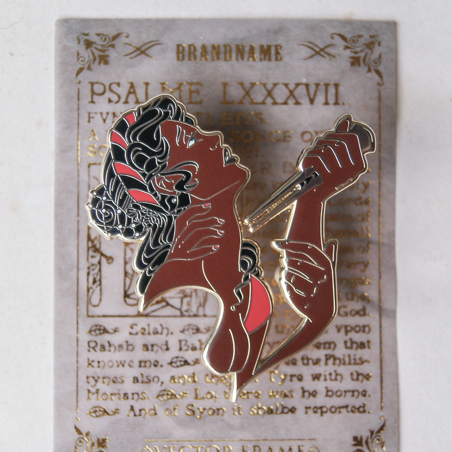 An enamel pin depicting a dark skinned woman with three hands holding a knife to her own neck. The pin is on top of a psalm.