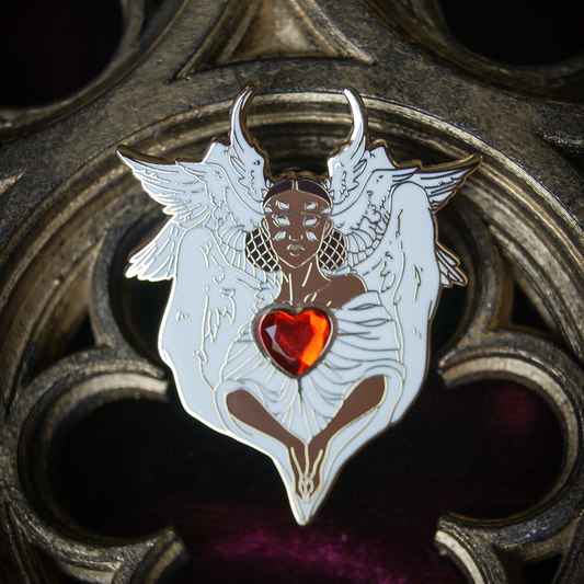 An enamel pin depicting a black angel with eight eyes and six wings. Her hands are clasped in prayer and there is a heart gem at her chest. 