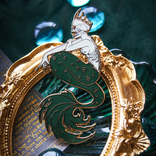 Photo of an enamel pin depicting a mermaid with long nails and a skull for a head. A spiked crown and ribcage corset complete her look. A gold frame sits behind. 