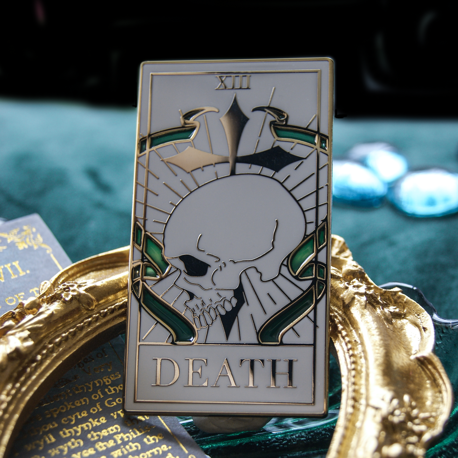 Enamel pin depicting the death tarot card, featuring stained glass in green. Below is a gold frame, in the background are green gems.