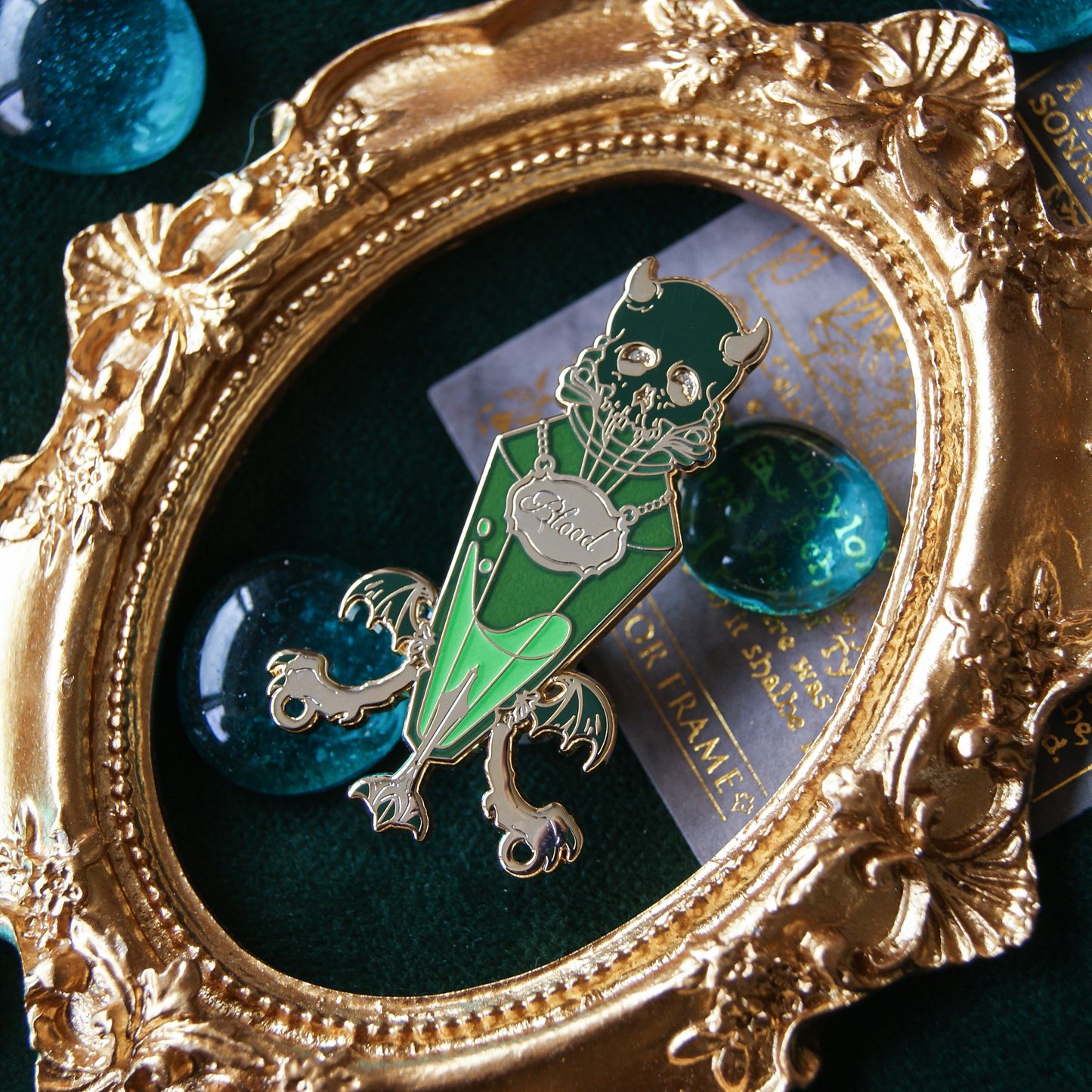 An enamel pin depicting a small vial filled with green liquid. a metal neckplate names this liquid as 'blood'. The stopper of the vial is a demon skull. In the background are green gems and a golden frame. 