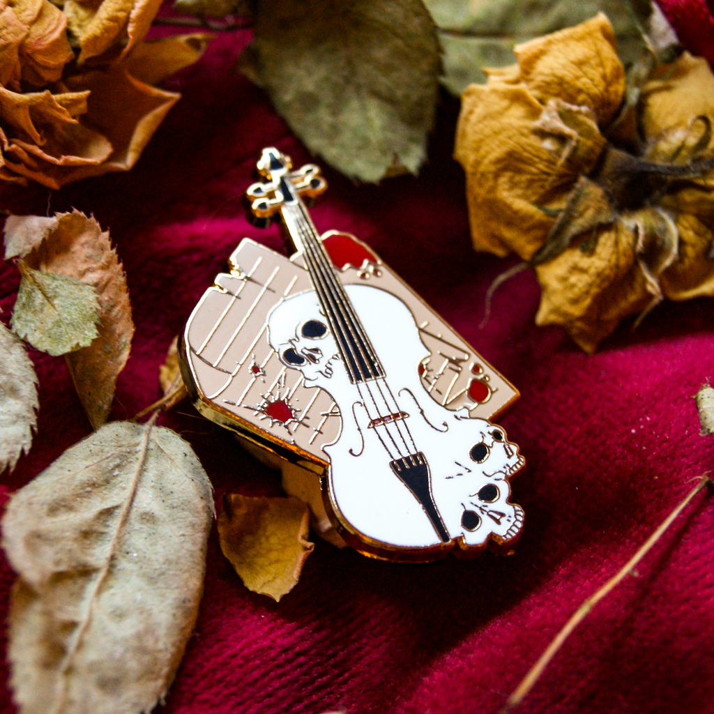 Enamel pin of a violin with skull design on a red fabric background with dried flowers and leaves.