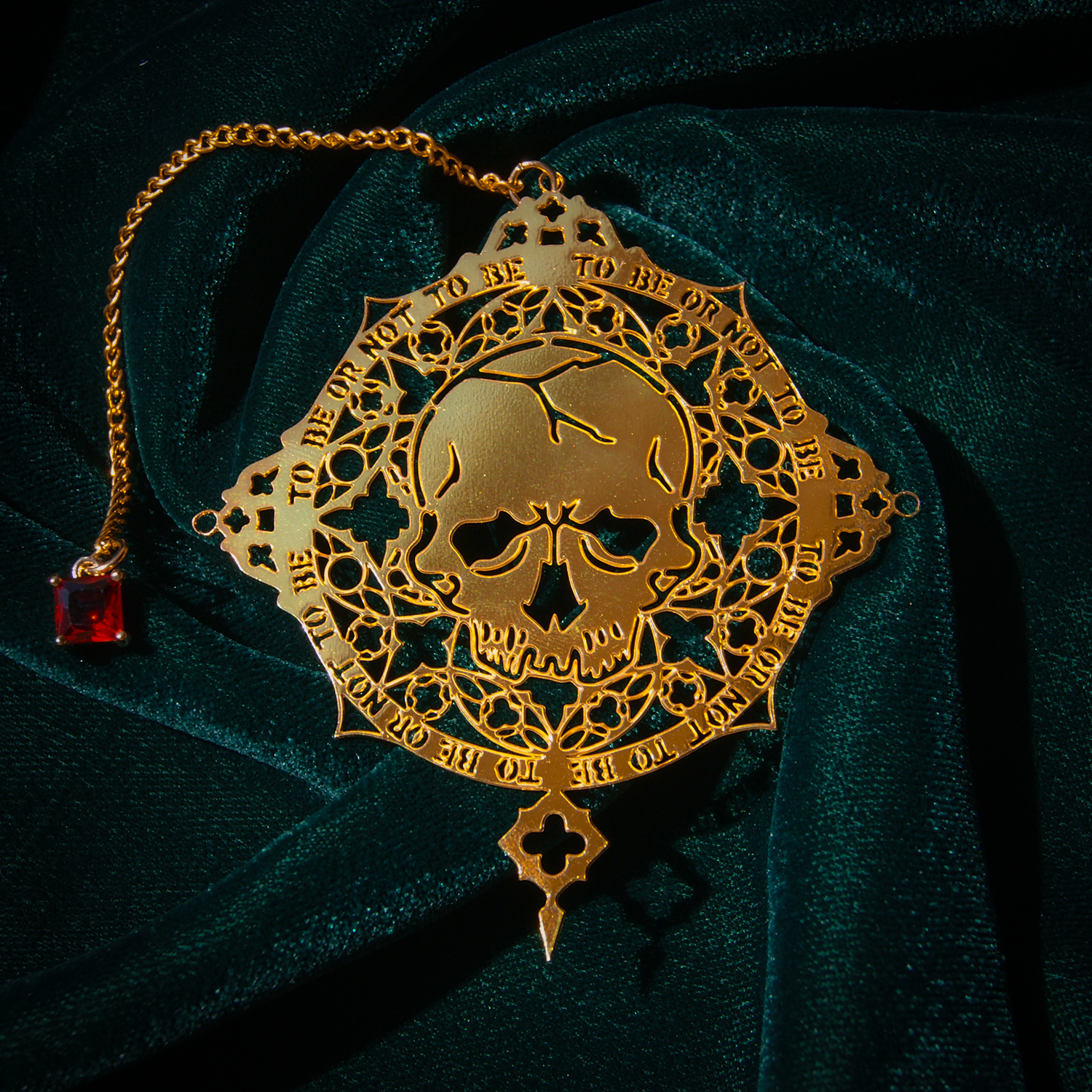 A metal bookmark in gold that has cut out sections to reveal a skull pattern. Around the edge reads To Be or Not to Be. A red gem is attached to the top by a gold chain. 