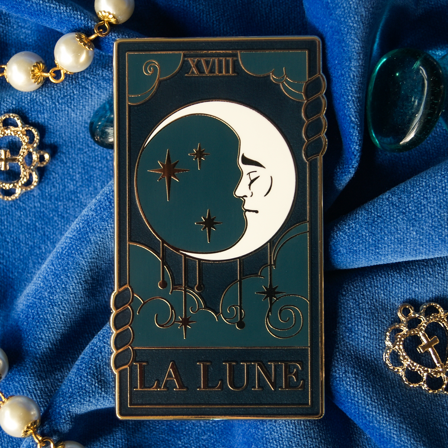 Photograph of an enamel pin depicting the moon tarot card. The pin is on tip of blue velvet scattered with pearls and decorations. 