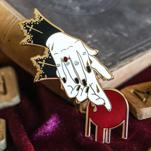 Enamel pin of two hands being washed. The hands have lace cuffs at the wrist and a red ring. At the fingertips is a red circle symbolising blood. Behind the pin is a book and some runes.