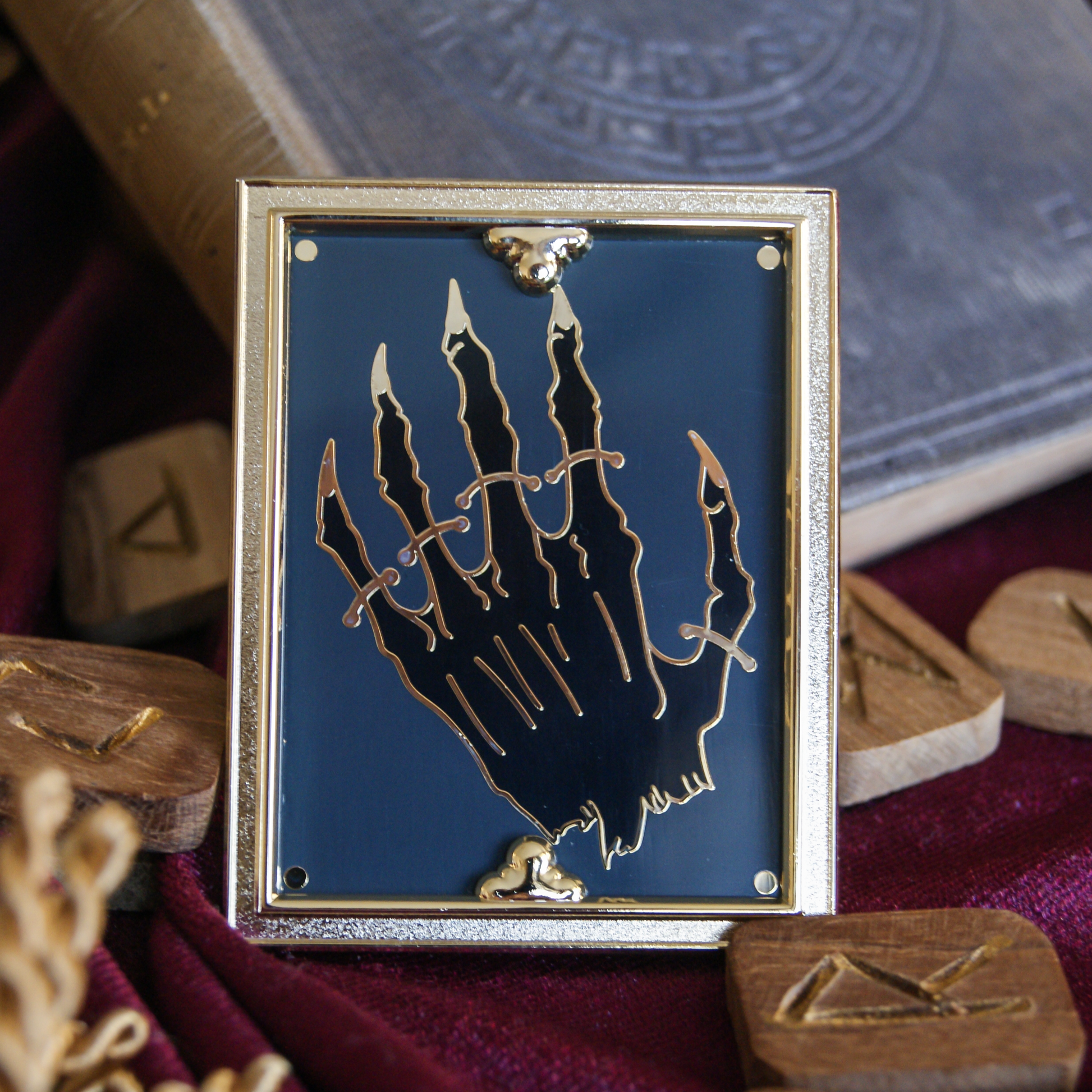 Photo of an enamel pin depicting a mummified hand strapped down like a museum exhibit. Around the hand is a 3D golden frame, and behind the pin is a book and some scattered wooden runes. 