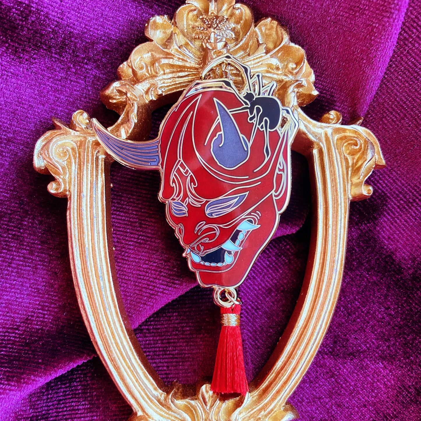 Photograph of an enamel pin depicting a Japanese oni mask being sat on by a black spider. Behind the pin is a golden frame and red velvet.