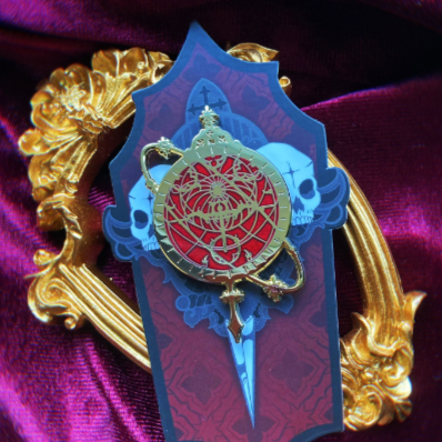 Photo of an enamel pin depicting an astrolabe with red background. Behind the pin is a backing card and a golden frame. 