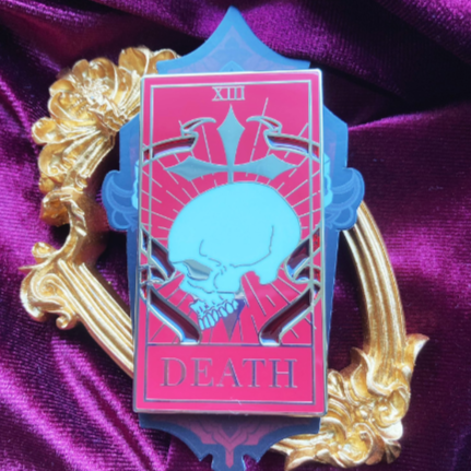 Photo of an enamel pin depicting a red colourway of the Death tarot card. Behind is a backing card and a golden frame. 