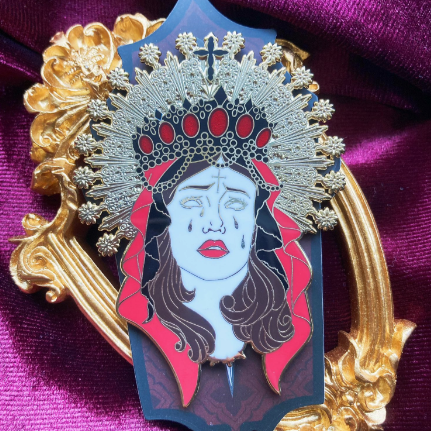 Enamel pin depicting the head of a woman wearing a crown and several veils. Around her head is a golden halo. She is crying. Behind the pin is a golden frame. 