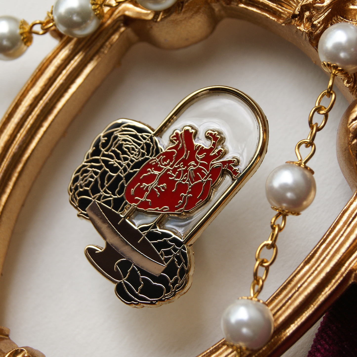 Enamel pin depicting a human heart inside a bell jar, backed by black peonies. Around the pin are pearls and a gold frame. 