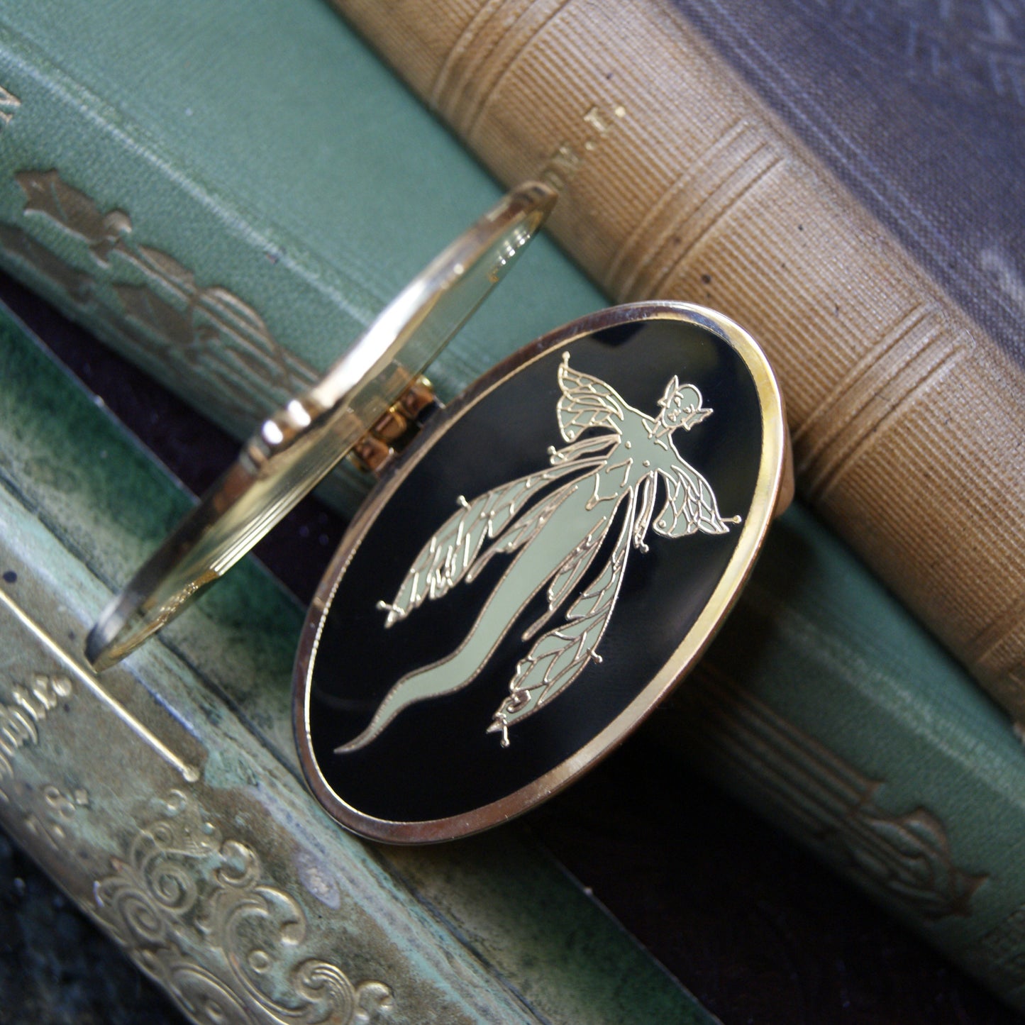 Photo of an enamel pin with a hinge; a river fairy is pinned underneath a glass cover. 