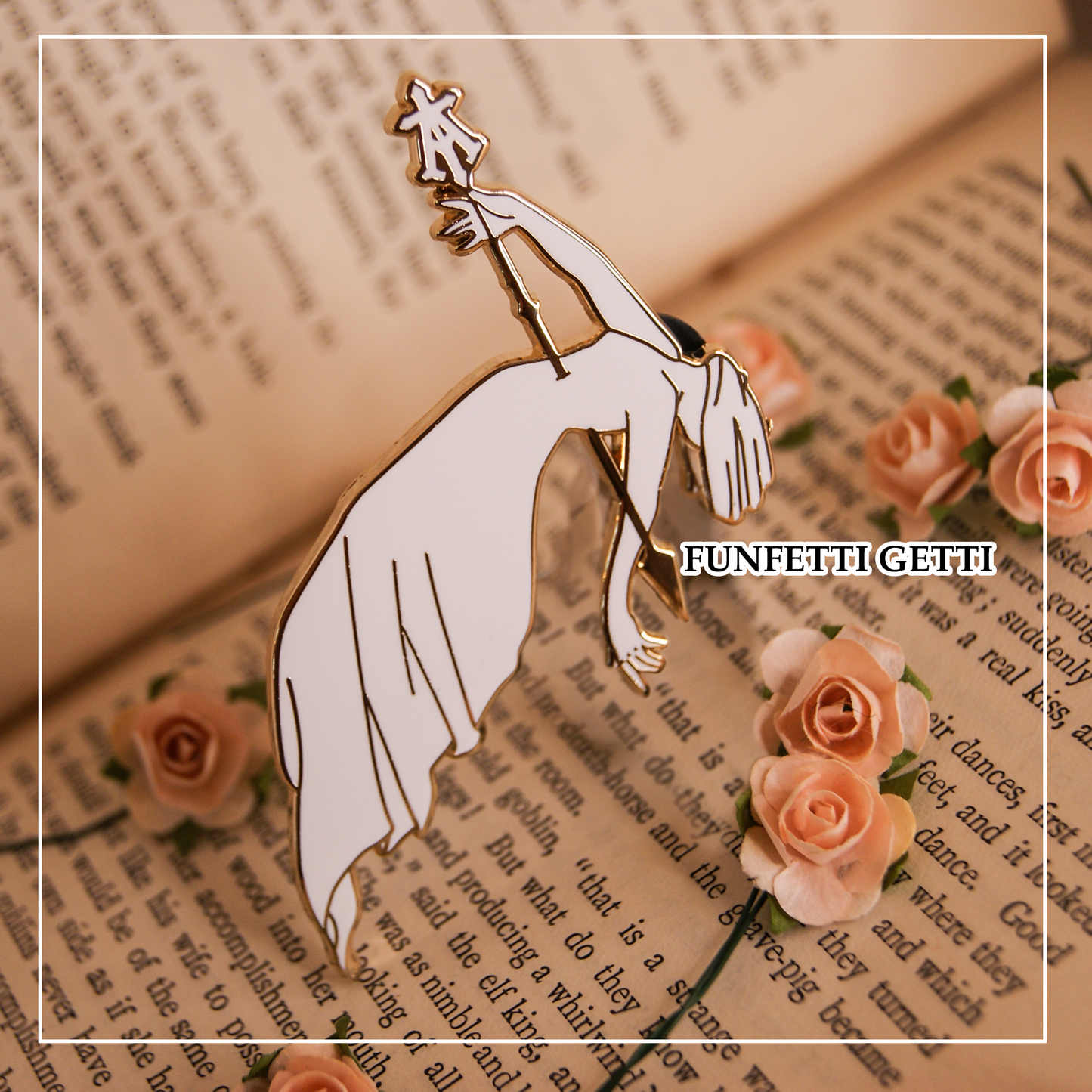 Photo of an enamel pin depicting a spear through the floating corpse of a woman in a long dress. A cloth is over her face. Behind the pin is a book and mini roses. 