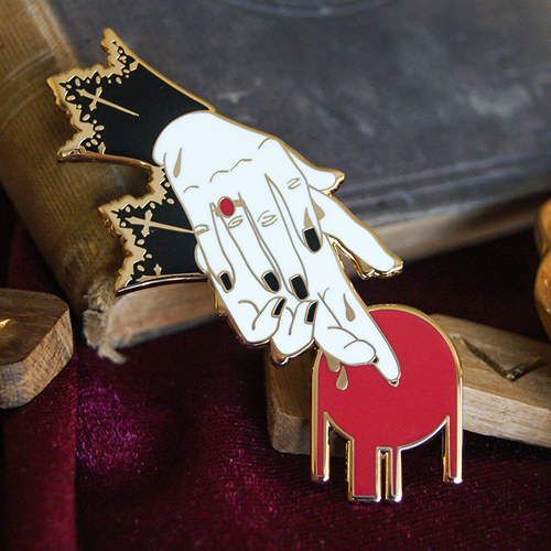 An enamel pin depicting two hands washing themselves. They have black lace cuffs and a ring. There is a red circle at the fingertips symbolising blood. 